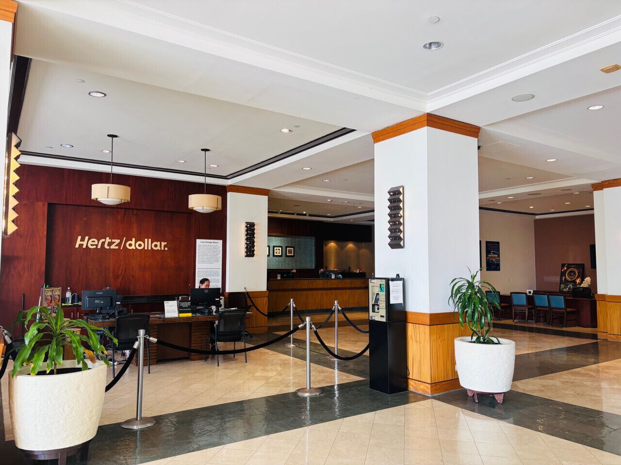 Un vestíbulo de hotel con una recepción, el logotipo de Hertz/un signo de dólar, y una planta grande en maceta.
