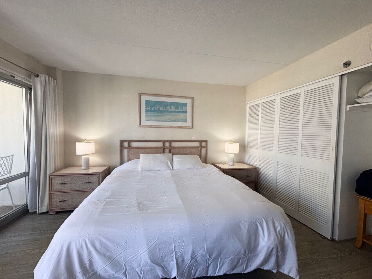 A hotel room with a large bed, two bedside tables with lamps, a window with curtains, a closet with louvered doors, and a framed picture on the wall.