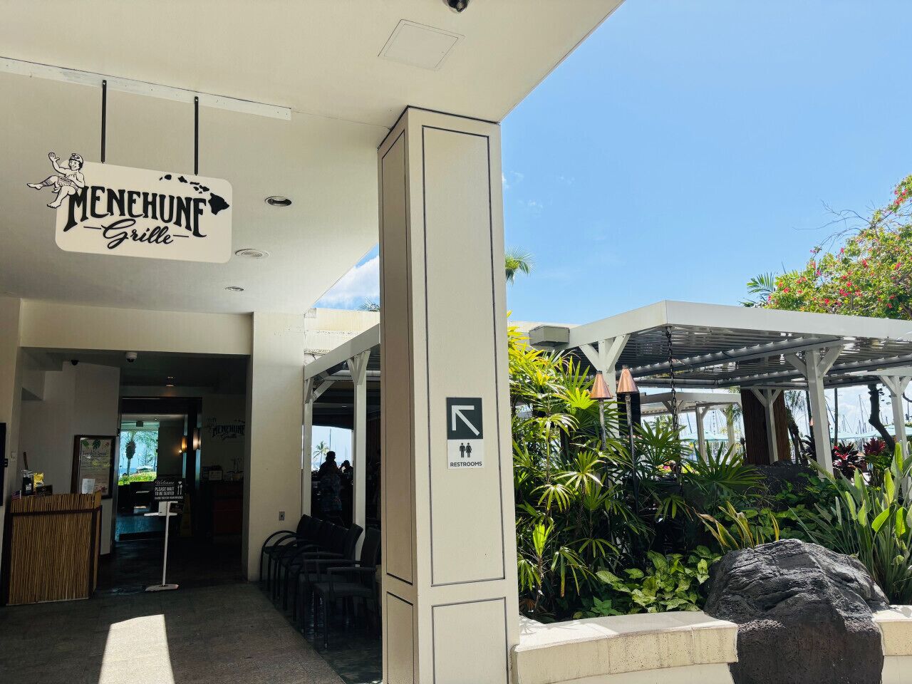 A restaurant entrance with a sign reading "MENENHUNE Grille," a signpost indicating restrooms, and a covered outdoor seating area with tropical plants.