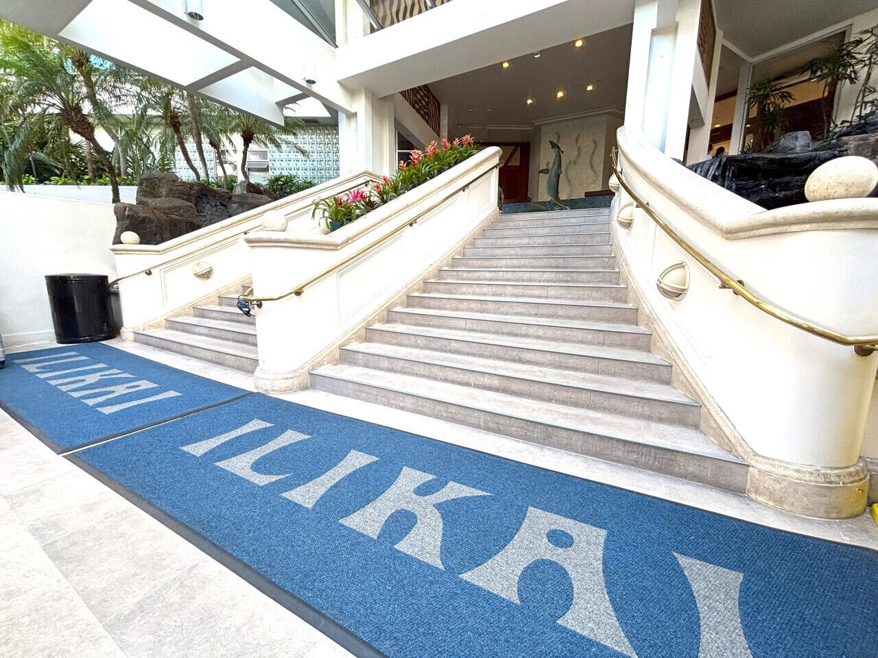 L’entrée d’un hôtel, avec un tapis bleu sur lequel est inscrit le mot « LIKAI », un escalier en marbre et une balustrade en pierre.