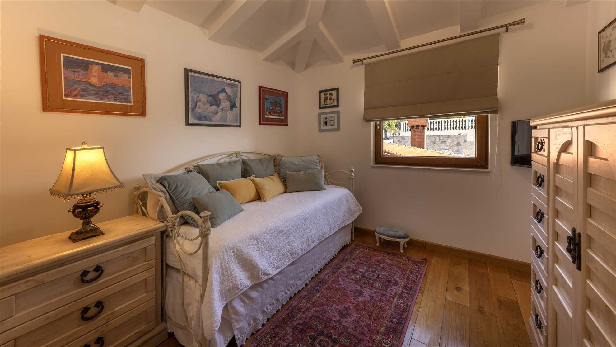 Ein Schlafzimmer mit einem Bett, einem Nachttisch, einer Lampe, einem kleinen Hocker, einem Fenster mit Blick und einem Holzschrank.