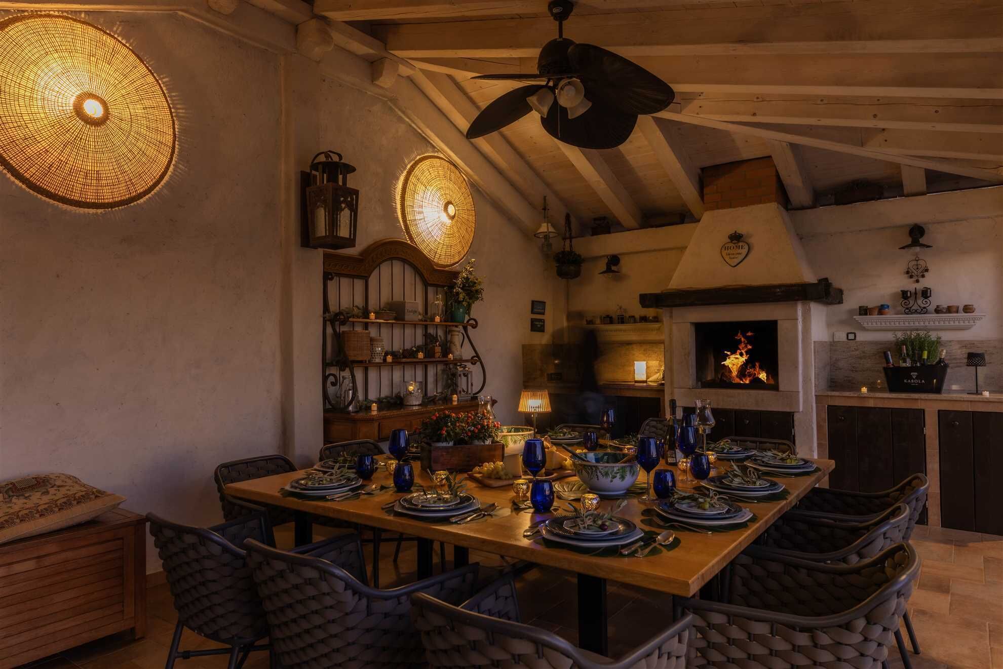 A dining area with a wooden table set for a meal, a ceiling fan, and a fireplace in the background.