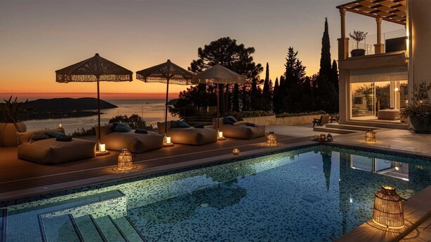 A poolside area with lounge chairs and umbrellas, a view of the sea at sunset, and a modern building with large windows.