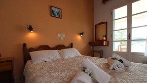 Una cama bien tendida con una colcha estampada, dos toallas enrolladas con motivos florales, una mesita de noche, una lámpara de pared y una ventana con vistas a la vegetación.
