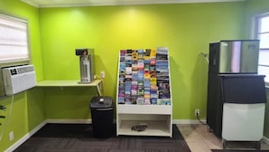 Une petite pièce aux murs verts, une étagère blanche avec des magazines, une poubelle noire, un climatiseur blanc et un réfrigérateur noir.