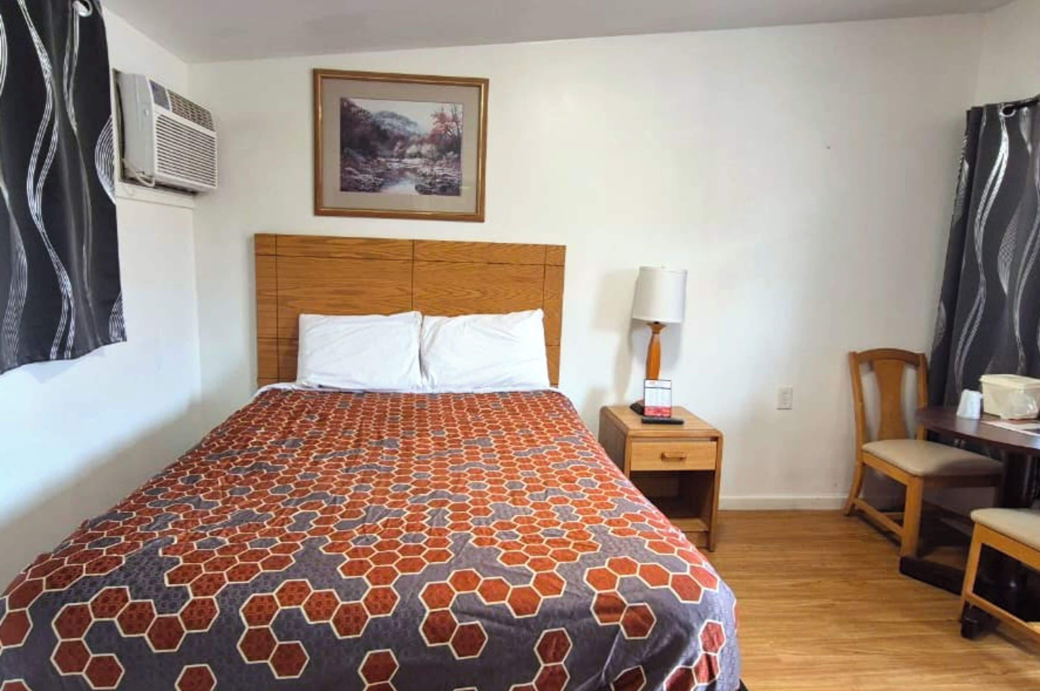 Una camera d'albergo con una testiera in legno, una coperta a motivi decorativi, un comodino con una lampada e una sedia.