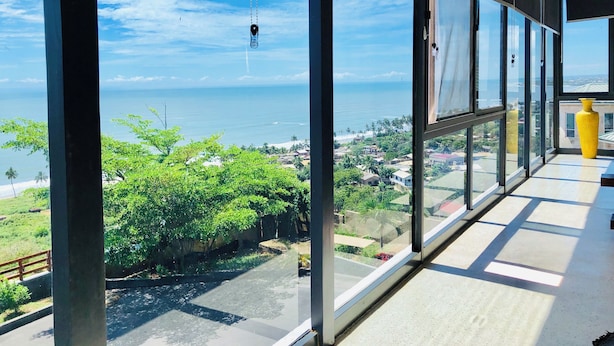 Un balcon doté de grandes fenêtres donnant sur une zone côtière, un vase jaune et un ciel dégagé.