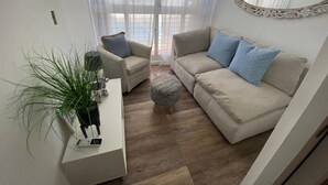 Una sala de estar con un sofá color beige, un banquillo redondo pequeño, una mesita auxiliar blanca con una planta y un espejo en la pared.