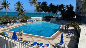Zona de piscina con vista despejada al océano, palmeras y un rascacielos al fondo.