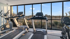 Un gimnasio con cintas de correr y bicicletas estáticas, ventanas amplias con vistas a una valla y palmeras.