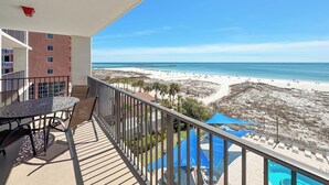 Un balcón con una mesa y sillas, con vista a una playa que tiene piscina y sombrillas.