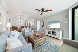 A living room with a sofa, coffee table, and TV. There is a ceiling fan and a kitchen in the background. - ORANGE BEACH RETREAT, GREAT VIEW OF THE PASS, GREAT AMENITIES, GREAT FOR FAMILY (Orange Beach)
