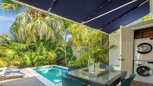 A poolside area with a table and chairs, a washing machine, and lush tropical plants. - HEMINGWAY'S HIDEOUT ~ Historic Old Town Cottage with Pool! (Key West)