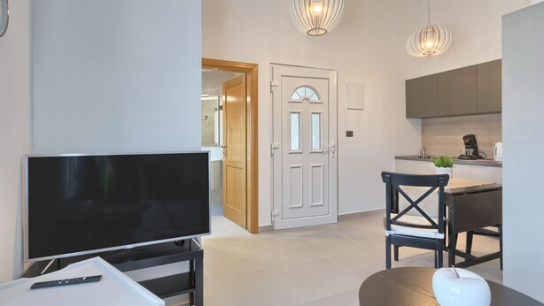 Un salon moderne avec un téléviseur à écran plat, une télécommande et un vase blanc posé sur une table. On y trouve également une chaise en bois foncé et une table à manger agrémentée d’un chemin de table noir. Deux lampes suspendues illuminent la pièce.