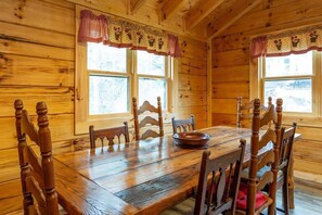 Une salle à manger en bois avec une grande table et des chaises, deux fenêtres avec des rideaux et une vue sur l’extérieur.