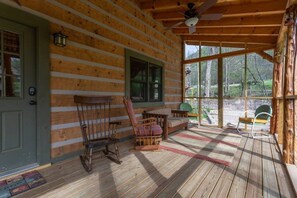 Une cabane en rondins avec un porche, un plancher en bois, un fauteuil à bascule, un banc et une vue sur l’extérieur.