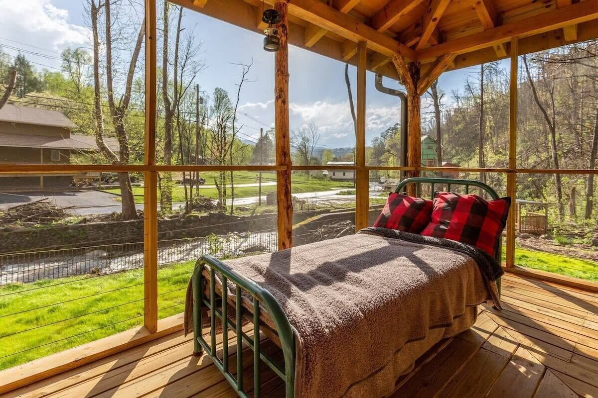 Een houten terras met een bed, grote ramen en uitzicht op een rivier en begroeiing.