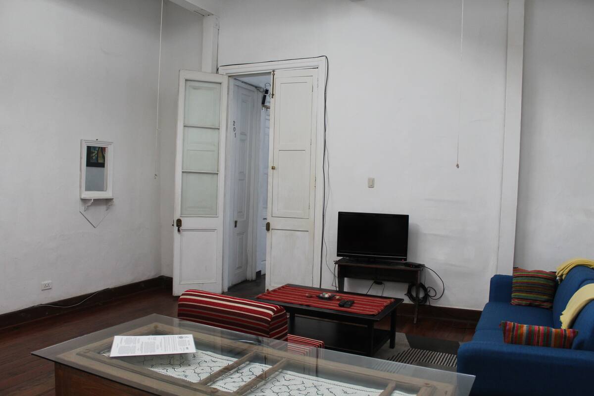 A living room with a glass coffee table, a sofa, a television, and a door leading to another room.