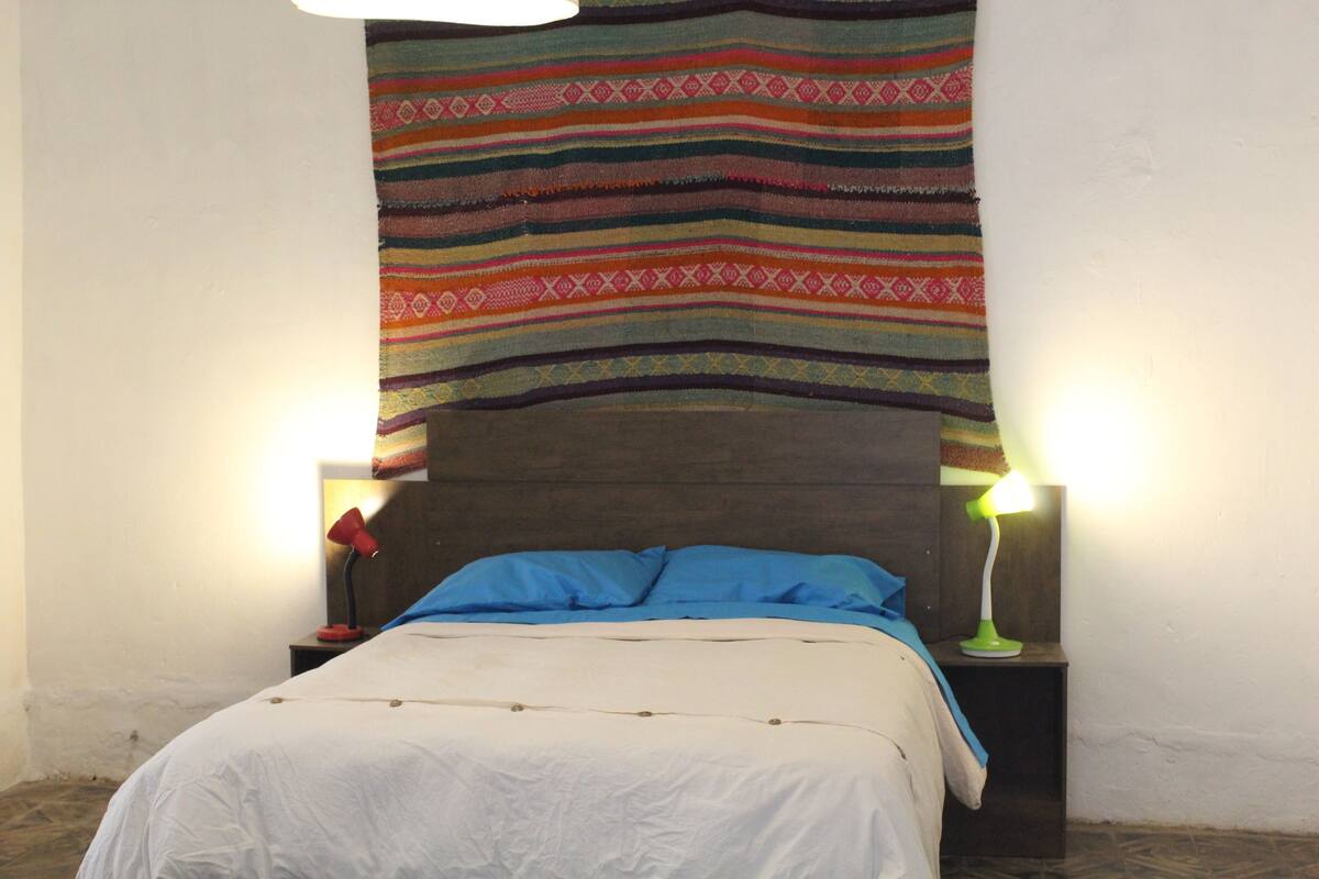 A bed with a white sheet and blue pillows, a colorful striped blanket, and two bedside lamps on wooden nightstands.