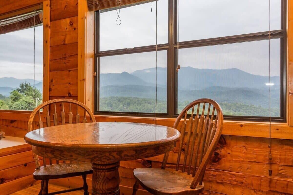Un intérieur en bois avec une table ronde et deux chaises, de grandes fenêtres offrant une vue sur la montagne.