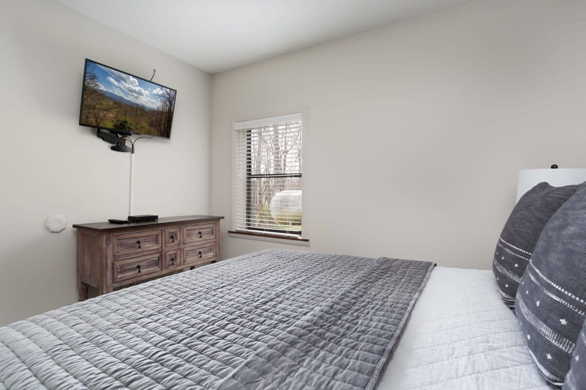 A bedroom with a bed, a dresser, a television mounted on the wall, and a window with blinds.