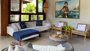 A spacious living room with a large window, a sofa, two armchairs, a round coffee table, and a painting of a person in water. - Retreat with Full Club and Cook (Praia do Forte)