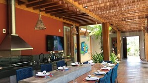 A long dining table with blue chairs, a kitchen with a large stove, and a wooden ceiling. - Retreat with Full Club and Cook (Praia do Forte)