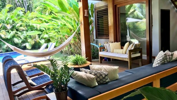 A wooden deck with a hammock, a pool, and a seating area with cushions and a table. - Retreat with Full Club and Cook (Praia do Forte)