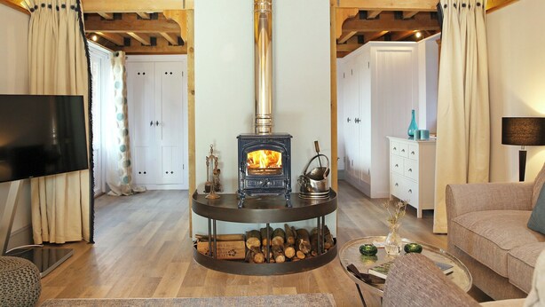 A cozy living room with a central wood-burning stove, wooden beams on the ceiling, a flat-screen TV, and a sofa. - The Barn at Banks Cottage (Pulborough)