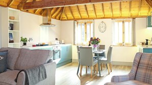 A cozy living room with a sofa and a plaid chair, a dining area with a table and chairs, and a kitchen with a window and a clock on the wall. - The Barn at Banks Cottage (Pulborough)
