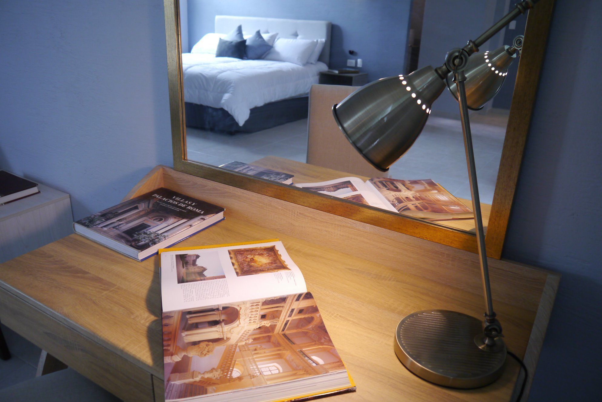 A hotel room with a wooden desk, a mirror reflecting a bed and a nightstand, and a desk lamp.