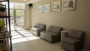 A living room with three grey sofas, a wooden coffee table, and a small round side table. There are framed pictures on the wall and a potted plant on the left side.