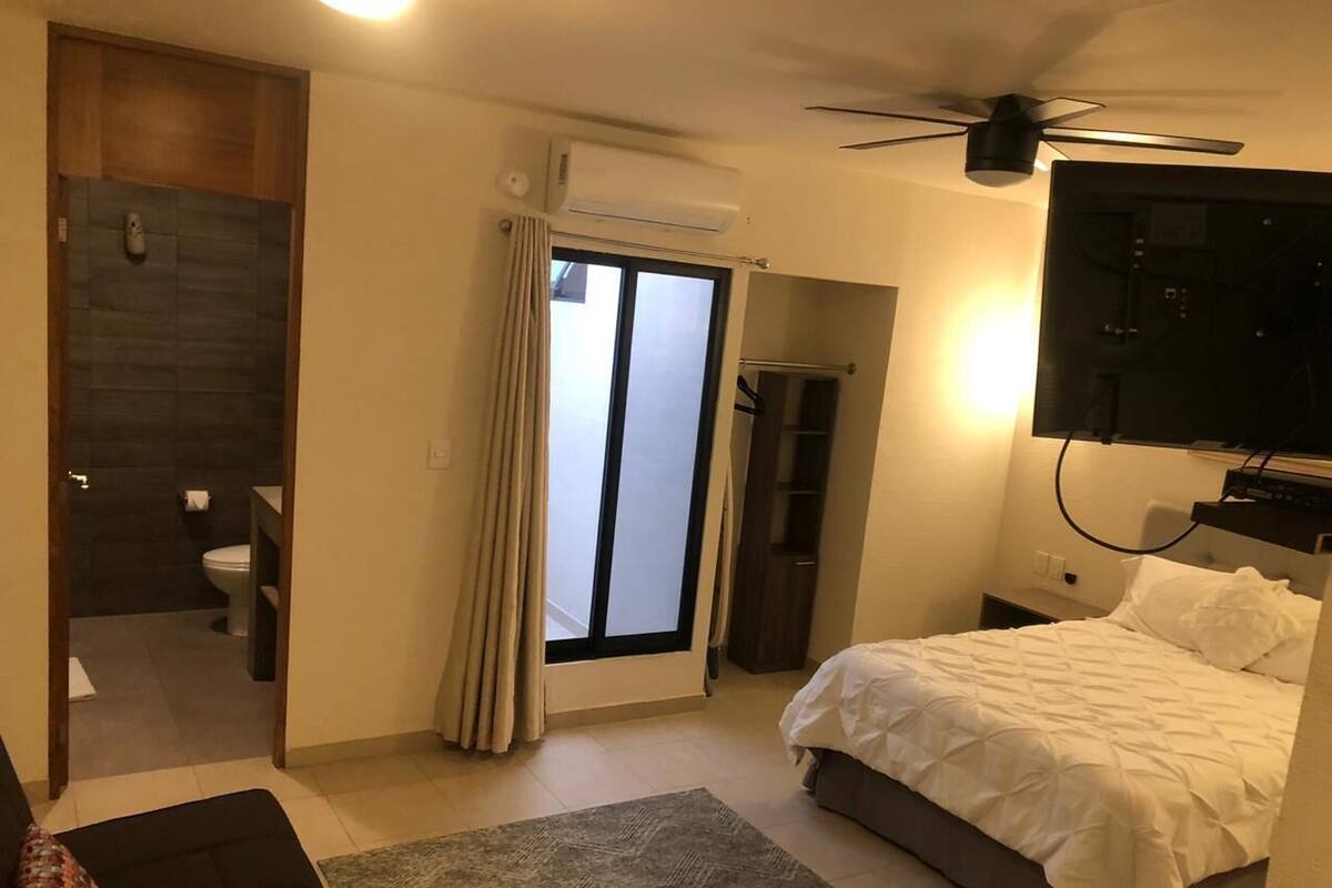 A bedroom with a bed, a TV, a ceiling fan, a window with curtains, and a bathroom visible through an open door.