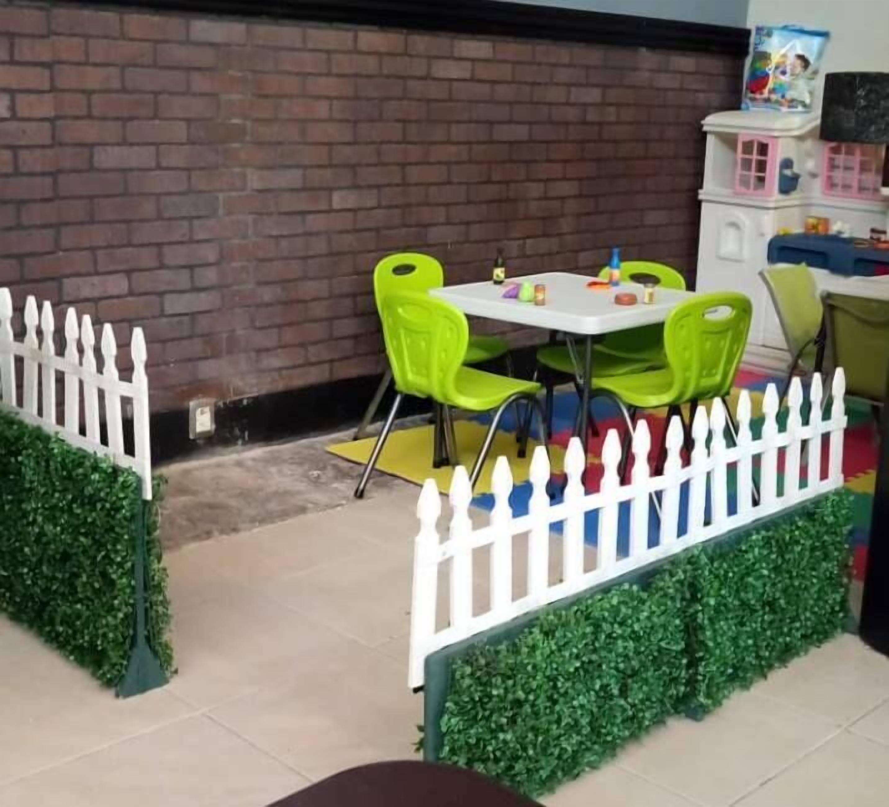 Un patio con pared de ladrillo, cerca blanca de piquetes y plantas artificiales verdes.