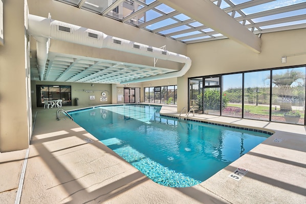 An indoor swimming pool with clear glass walls, a ceiling with skylights, and a poolside area with a patio.