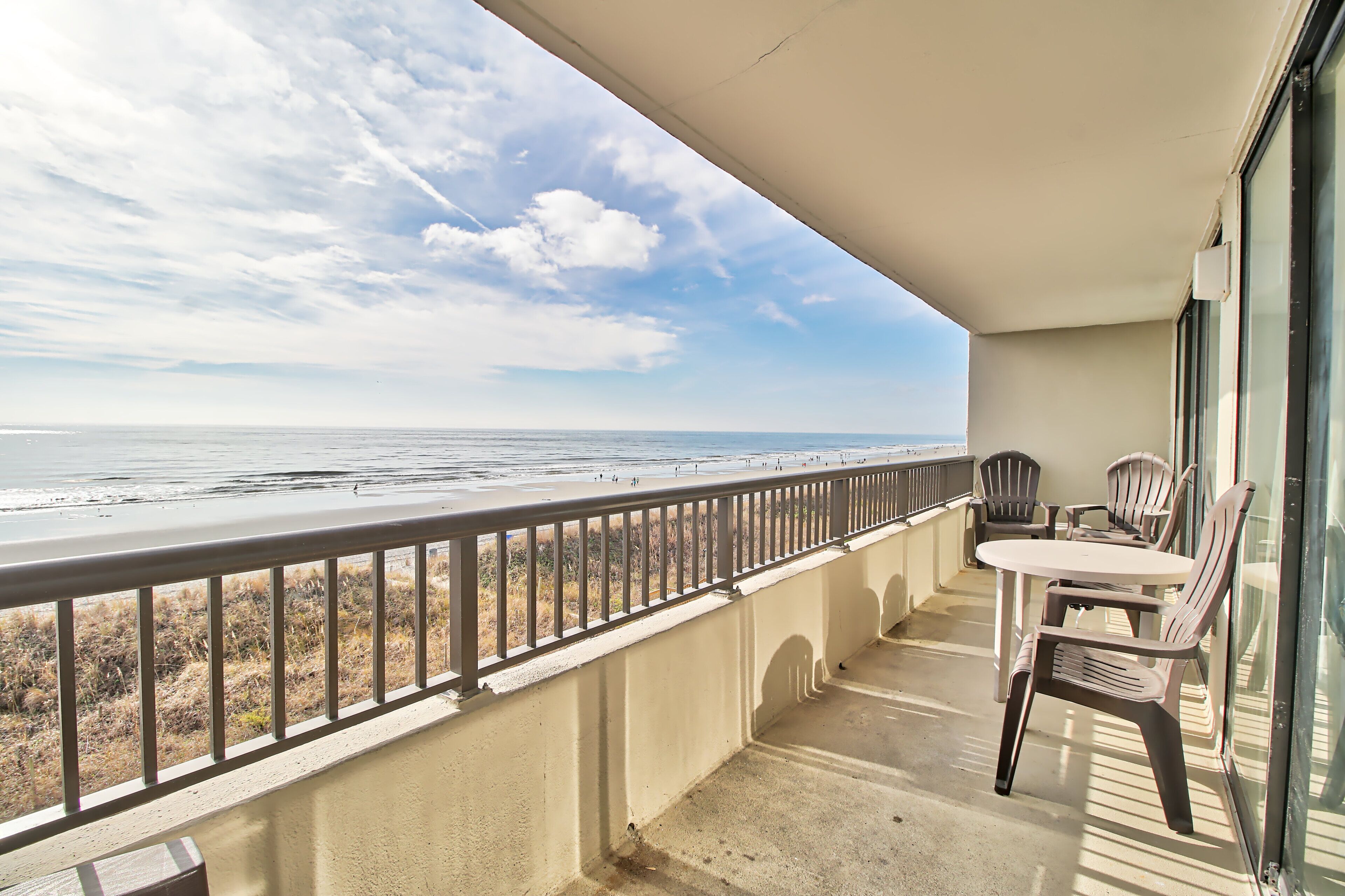 A balcony with a view of the beach, a table, and two chairs.
