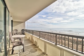A balcony with a view of the beach, a table, chairs, and a glass door.