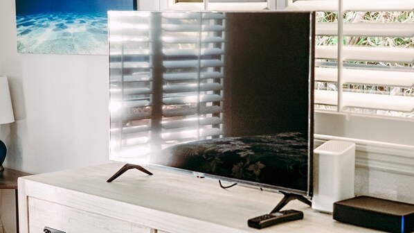 Ein Flachbildfernseher auf einem Ständer mit Fernbedienung, vor einem Fenster mit Jalousien platziert.