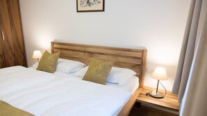 Ein ordentlich bezogenes Bett mit weißen Leinen und zwei Kissen, ein hölzernes Kopfteil sowie je eine Nachttischlampe auf Holz-Nachttischen.