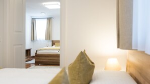 A hotel room with a bed, a bedside table, a lamp, and a view of another room with a bed and a window.