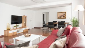 A modern living room with a red sofa, a white coffee table, and a TV on a wooden stand. There is a dining area with a table and chairs in the background.