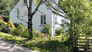 A white house with a sloped roof, surrounded by lush greenery and a wooden gate. - 2 bedroom accommodation in Arduaine, near Oban (Arduaine, near Oban)