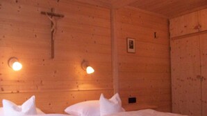 Een houten kamer met een kruis aan de muur, twee bedden met witte lakens en wandlampen.