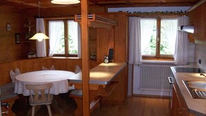 Een houten blokhut van binnen met een eettafel, stoelen, een keukenhoek en een raam met gordijnen.