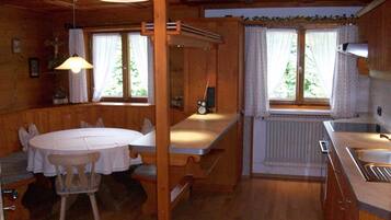 A wooden cabin interior with a dining table, chairs, a kitchen area, and a window with curtains.