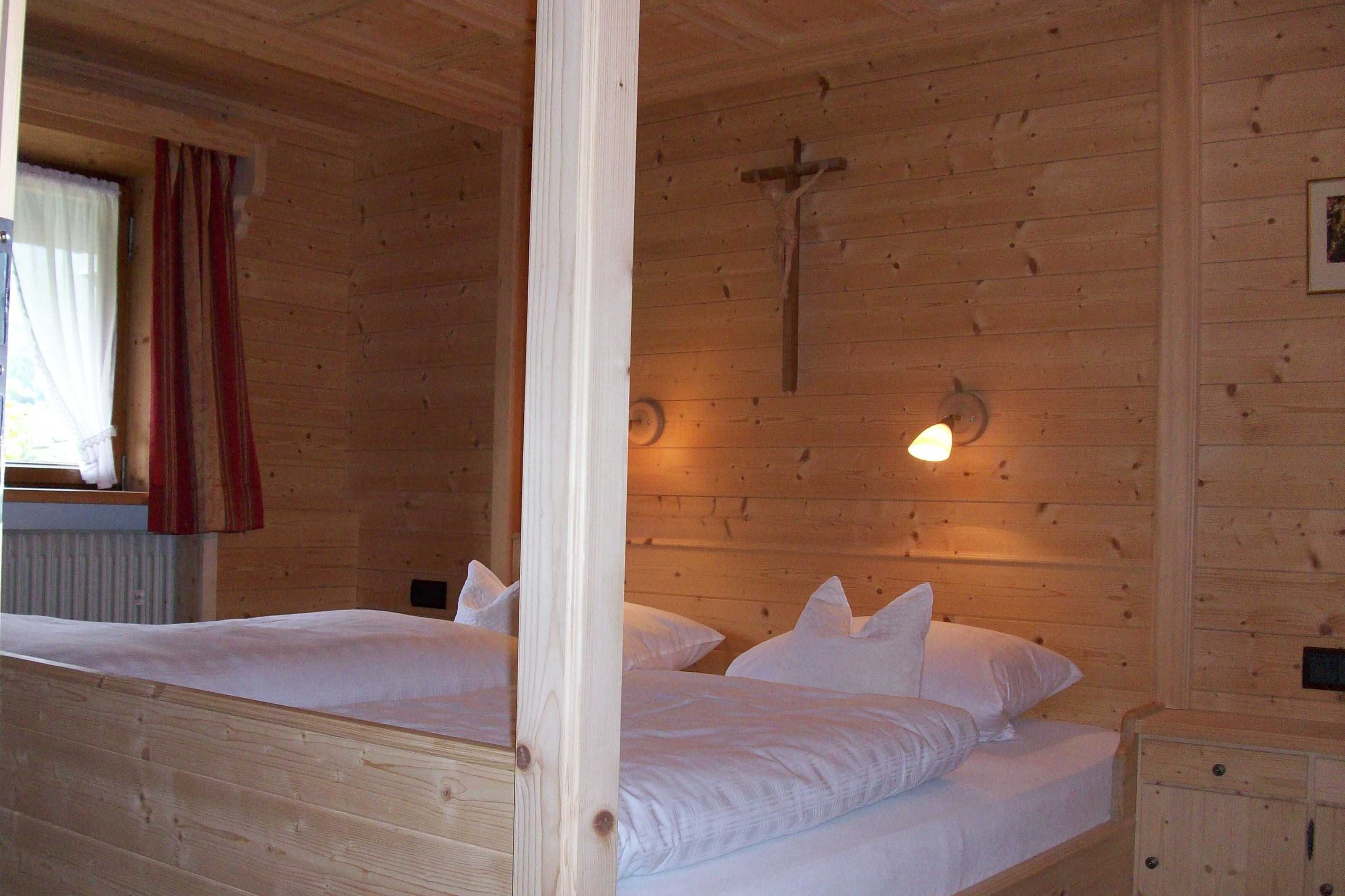 A wooden cabin room with two beds, a wooden cross on the wall, and a window with curtains.