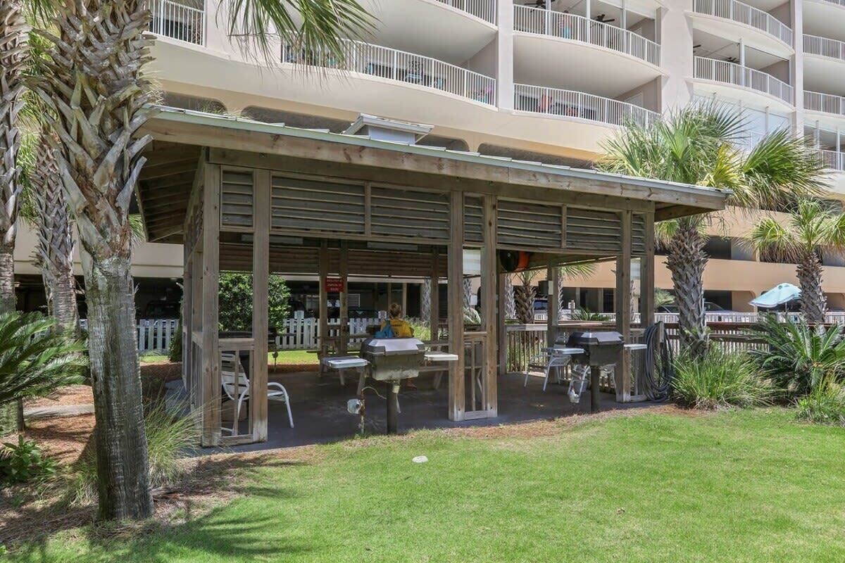A covered outdoor seating area with tables and chairs, surrounded by palm trees and a building with multiple balconies in the background.