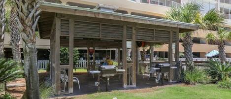 A covered outdoor seating area with tables and chairs, surrounded by palm trees and a building with multiple balconies in the background.