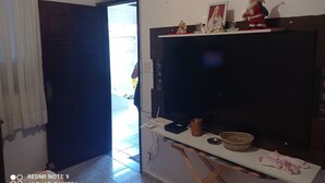 A room with a large TV on a stand, a door leading outside, and a table with various items including a basket and a small statue. - Family home (Sítio do Campo)