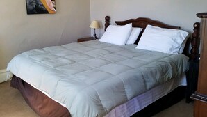 Un lit bien fait, avec une couette de couleur claire, une tête de lit en bois et une table de chevet surmontée d’une lampe. Un tableau est accroché au mur, au-dessus du lit.